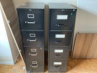 Photo showing two black metal file cabinets standing side by side. Each cabinet has four drawers with metal handles and label holders.