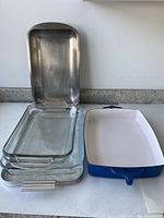 Four nested clear glass rectangular baking dishes with metal handles and one blue and white rectangular roasting tray placed side by side.