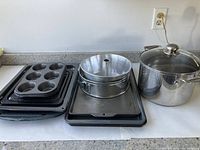 Photo showing an assortment of baking trays including muffin tray, sheet pans, springform cake pan, loaf pan, and a stainless steel pot with glass lid on a countertop.