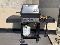 Front view of black Broil King propane BBQ grill with closed lid, two side shelves, propane tank underneath, and grilling tools on the right shelf.