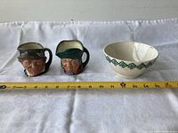 Front view of two character mugs and one decorative bowl placed on white fabric with measuring tape showing size.
