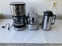 Photo showing Oster coffee maker on the left, Cuisinart food processor in the center, and stainless steel vacuum carafe on the right on a white countertop.