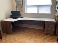 Front view of the L-shaped desk under a window with a computer monitor, keyboard, and mouse on the surface (computer not included).
