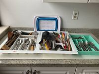Photo showing the organized white trays holding an assortment of metal forks, spoons, knives, and various kitchen utensils including peelers, ladles, wooden and plastic handled tools on a granite countertop with a collapsible cutting board behind.