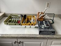Photo showing assorted cutlery in green and yellow trays, kitchen knives in wooden block with white handles, a boxed corkscrew, and various kitchen utensils and tools on a counter.