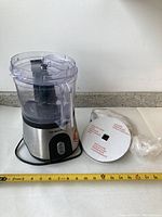 Hamilton Beach food processor with clear plastic bowl, lid, stainless steel base, and black blade inside bowl. Instruction manual and sealed accessory included. Tape measure for size reference.