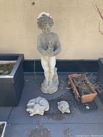 Garden sculptures displayed on an outdoor patio surface against a light wall, including a tall child figure statue and two smaller animal figures near the base, alongside large black planters and a rectangular terracotta planter containing dried plant remains.