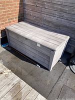 Closed garden storage box viewed from the side against wooden fence and brick wall outdoors.