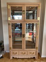 Front view of wooden glass display hutch with brass hinges, latches, and three drawers at base.