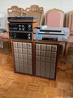 Full view of the stack of Hi-Fi components and speakers, with two vintage speakers in front on the floor, amplifiers and media players stacked higher on a table, and several upholstered chairs in the background.