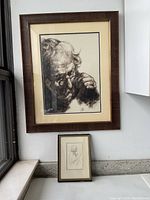 Photo shows two framed drawings on a windowsill: a larger, detailed pencil drawing of an elderly man wearing glasses and resting his head on his hand, and a smaller pencil sketch of a bearded elderly man.