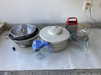 Photo of kitchen utensils on table including metal colander, white salad spinner, glass measuring cups, blue lidded glass containers, and metal box grater with red handle.