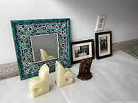 Photo showing ceramic tile mirror, two white onyx/alabaster bookends, two small framed black and white photos, and a small carved wooden sculpture on white paper on a countertop.