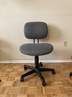 Front view of a gray wheeled office chair with fabric upholstery and black five-wheel base on a parquet floor.