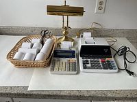 Photo of office lot items including two calculators, basket of paper rolls and scissors, and brass desk lamp on white surface against white wall.