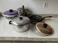Photo showing five pieces of cookware arranged on countertop including enamel pot, stainless steel stockpot, aluminum pot, small frying pan, and copper-colored lid