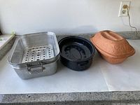 Photo showing three roasting pans: aluminum rectangular pan with rack, black speckled oval pan, and terracotta clay baker with lid.