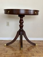 Front view of round wooden table showing turned pedestal base, three curved legs with brass feet, two brass ring handle drawer fronts.