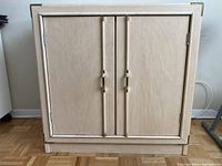 Closed blond wood cabinet showing two doors with brass handles and hinges.