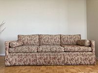 Front view of the vintage sofa fully upholstered with floral fabric showing three seat cushions, three back cushions, two bolsters, and full skirt covering the base.