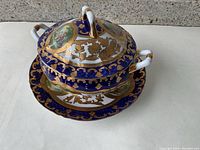 View of the top and side of the ornate porcelain tureen on attached plate, featuring blue and gold decoration with painted scenic ovals and gold accents.