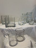 View of all 8 assorted clear glass vases in various shapes and sizes on a white cloth background.