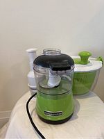 KitchenAid food processor in front, Cuisinart salad spinner at back right, and manual chopper at back left on white cloth surface.