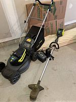 Push mower and string trimmer side by side with battery and charger on concrete floor