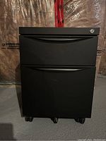 Front view of the black 2-drawer file cabinet with wheels, showing the closed drawers and caster wheels at the bottom.