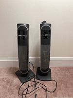 Two black and silver Bionaire electric tower heaters side by side, showing full height and bases with power cords in front.