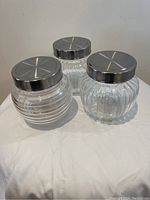 Three clear glass jars with decorative ridged patterns on glass and silver metal lids arranged on white cloth.