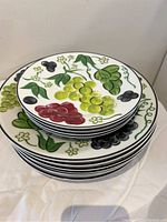 Stack of 4 small dessert plates featuring grape cluster designs in red, green, and black, with green leaves and small yellow flowers on a white background with black rim.