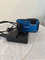 Front view of blue Panasonic Lumix digital camera with lens and brand logo, with battery charger in foreground.