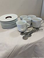 Overview of all items: 5 white espresso cups, 6 white saucers, 5 metal stir spoons arranged on a white surface.