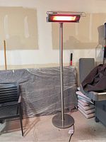 Full view of electric outdoor standing heater with glowing heating element, stainless steel pole, and round base.