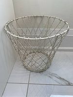 White wired metal waste bin on tiled floor against wall, showing full round shape and sturdy construction.