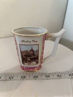 Side view of small porcelain vase showing scenic Karlovy Vary image, gold trim and handle detail next to ruler for scale.