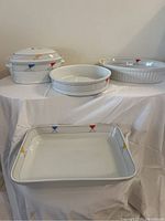Four-piece ceramic bake and serve set displayed on white cloth background. Includes 2 round covered casseroles, 1 oval dish, 1 rectangular dish. All pieces white with colorful geometric flag motifs.