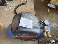 Blue electric air compressor with pressure gauge and dual hose connectors visible from above. Black motor cover and power cord attached.