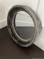 Image showing the front and side view of the 16 inch round mirror with black antiqued distressed frame against a white wall and floor.