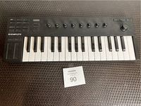 Black Native Instruments Komplete MIDI keyboard controller with 25 keys, control knobs, buttons, and joystick on textured surface with maxsold number 90 tag.