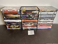 Photo showing three stacks of movie DVDs, approximately 44 DVDs total, with readable titles including The Goonies, Black Hawk Down, Saw, Ghostbusters, Happy Gilmore, and more.