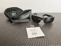 Oho Sunshine black camera sunglasses, zipped hard case, USB charging cable, and lot number card on textured surface
