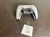 Single photo of the PlayStation 5 DualSense wireless controller placed on a textured dark surface with a MaxSold auction bid ticket showing lot number 125.