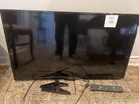 Front view of a Seiki smart TV with black screen, two black detachable legs and a remote placed in front on a carpet.
