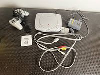 Photo of Sony PlayStation One console, two original controllers, power adapter, and composite AV cables arranged on a textured surface.