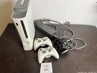 White Xbox 360 console standing vertically with two white and one black wireless controllers in front. Power adapter placed behind controllers. Photo shows full lot contents with minor surface wear visible.