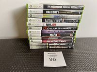 Stack of 12 Xbox 360 video game cases against a wall on a textured floor mat. Visible titles include popular games like The Walking Dead Survival Instinct, Call of Duty Advanced Warfare, and Grand Theft Auto IV.