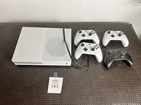 Top view of white Xbox One S console with power cable beside three white and one black wireless controllers on dark surface, lot label '143' visible.