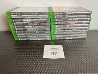 Photo showing the spines of 21 Xbox One game cases stacked in two piles on a black woven surface along with a MaxSold lot card 97.